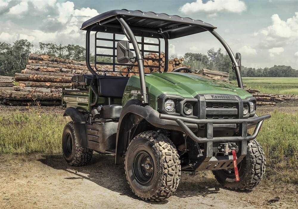 Green Kawasaki Mule utility vehicle parked outdoors in front of stacked logs under a cloudy sky with protective rollover cage and rugged tires.