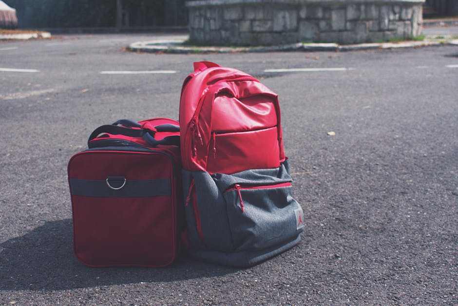 Two red UTV storage bags on asphalt, including a backpack and a rectangular bag with zipper compartments.