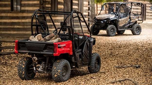 Red Honda Pioneer 520 kids side by side UTV with cargo bed loaded with firewood, parked on woodchip ground near a wooden fence with another black UTV in background, showcasing kids side by side models.