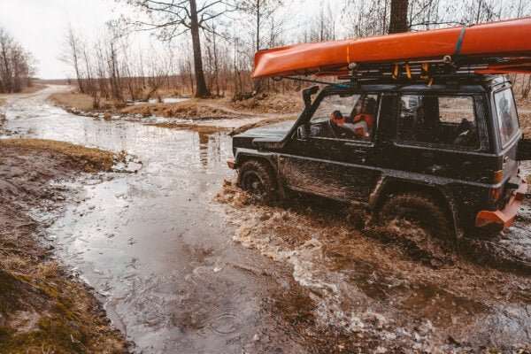 Black off-road car with kayak on roof driving through muddy water trail in forest area