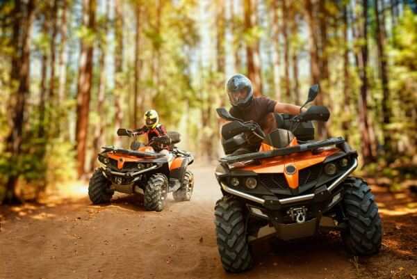 Two riders on orange ATVs riding through a sunlit forest trail, showcasing outdoor adventure and ATV accessories.