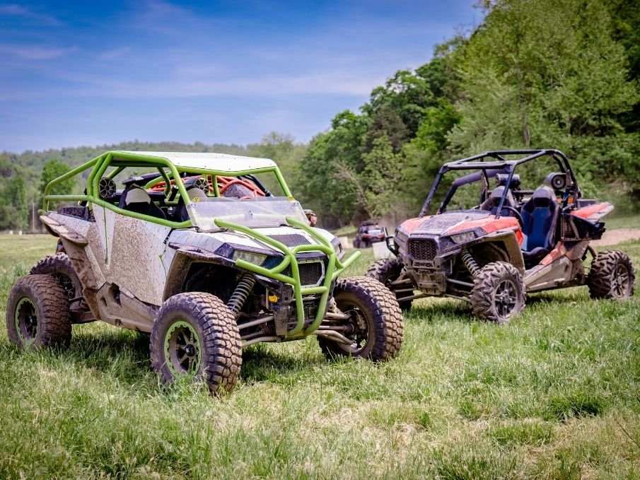 Two utility terrain vehicles (UTVs) parked on a grassy field with trees and blue sky in the background