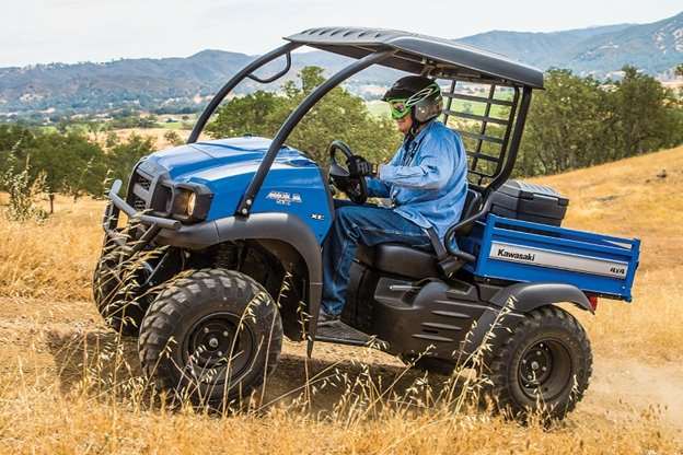 Blue Kawasaki Mule SX XC UTV being driven on rough terrain for farm work with a rider wearing protective gear and helmet.